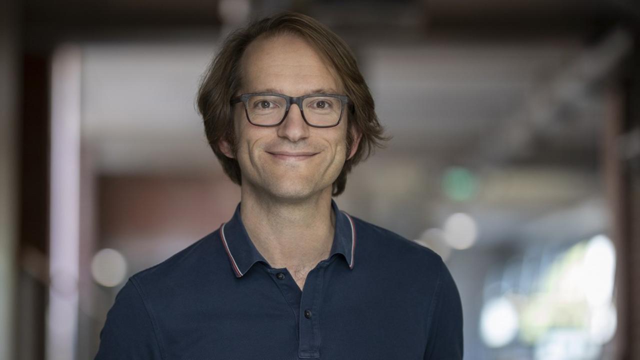 Ein jugendlich wirkender Mann in dunkelblauem Poloshirt. Er trägt eine dunkle Hornbrille und mittellanges gescheiteltes Haar. Ein jugendlich wirkender Mann in dunkelblauem Poloshirt. Er trägt eine dunkle Hornbrille und mittellanges gescheiteltes Haar.