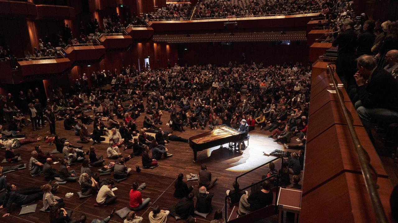Zuschauer sitzen und liegen in einem Konzertsaal, in der Mitte sitzt ein Pianist an einem Flügel Zuschauer sitzen und liegen in einem Konzertsaal, in der Mitte sitzt ein Pianist an einem Flügel