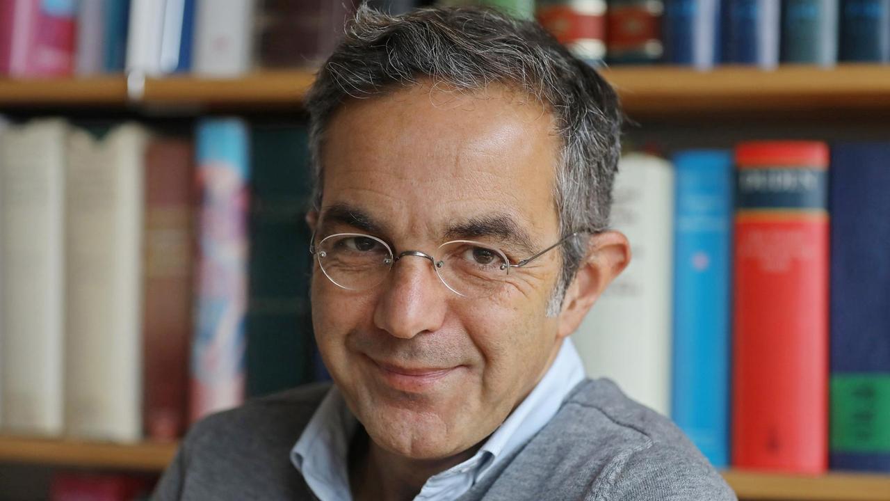 Navid Kermani, Schriftsteller, blickt lächelnd in die Kamera, im Hintergrund ist ein Bücherregal. Navid Kermani, Schriftsteller, blickt lächelnd in die Kamera, im Hintergrund ist ein Bücherregal.