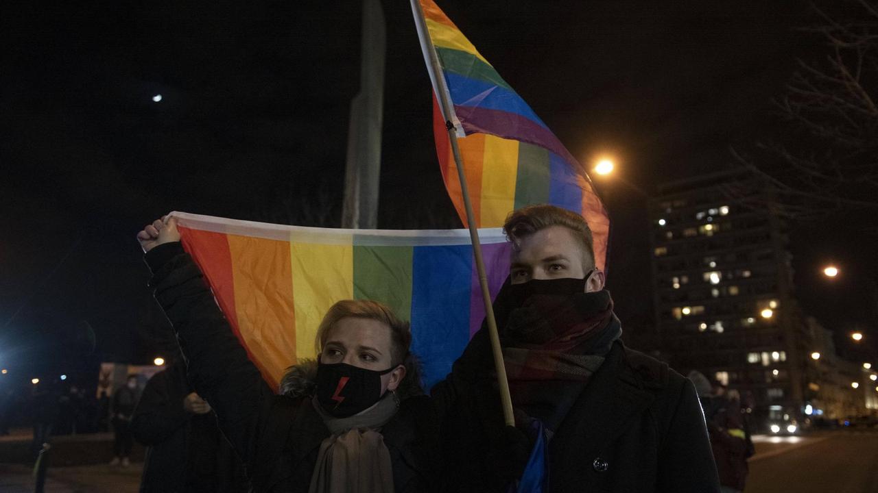 Zwei dunkel gekleidete jugen Menschen halten vor nächtlichem Himmel Regenbogenflaggen hoch. Zwei dunkel gekleidete jugen Menschen halten vor nächtlichem Himmel Regenbogenflaggen hoch.