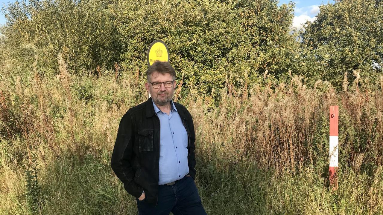 Ein Mann mit schwarzer Jacke und Hose und hellblauem Hemd, steht - die Hand in der Tasche - in einer grünen Landschaft. Ein Mann mit schwarzer Jacke und Hose und hellblauem Hemd, steht - die Hand in der Tasche - in einer grünen Landschaft.