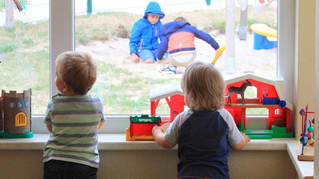 Zwei Kinder stehen am Fenster einer Kita und gucken anderen Kindern draußen beim Spielen zun Zwei Kinder stehen am Fenster einer Kita und gucken anderen Kindern draußen beim Spielen zun