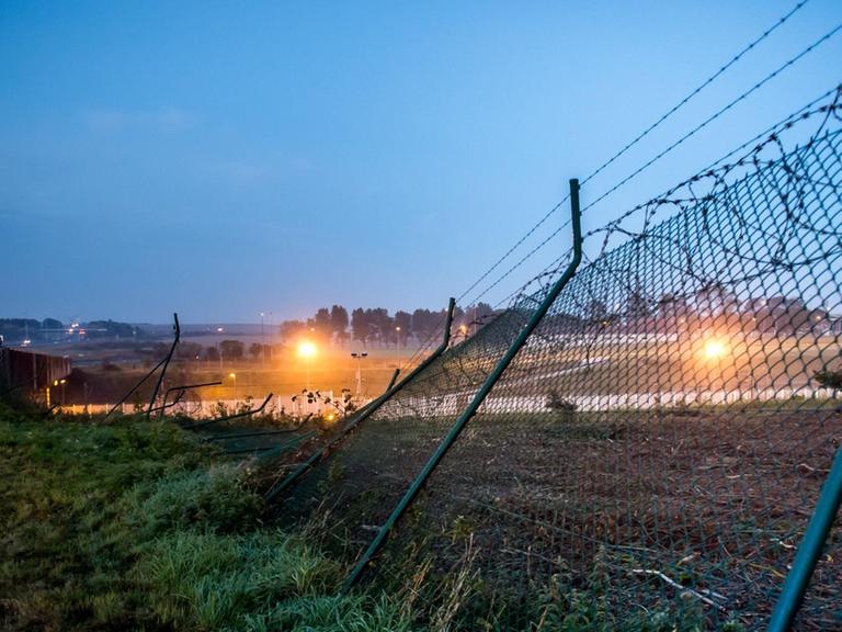 Man sieht den auf den Boden gebogenen Zaun im Morgengrauen; er wird von einem Scheinwerfer aus dem Eurotunnel-Gelände angestrahlt.