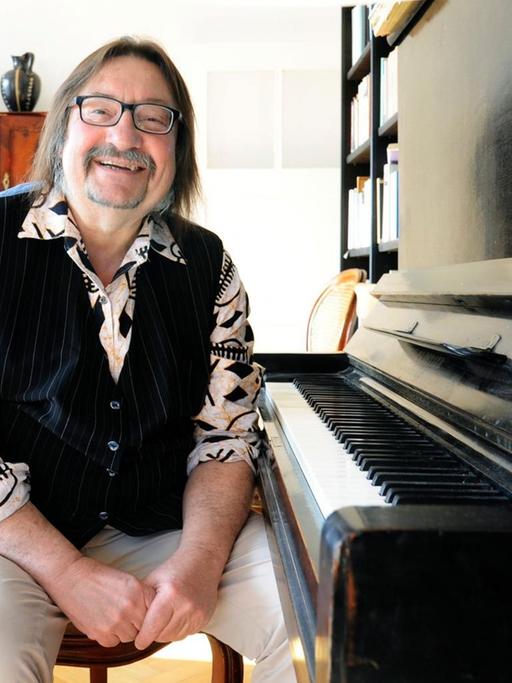 Manfred Maurenbrecher sitzt in seiner Wohnung am Klavier und schaut freundlich in Richtung Kamera.