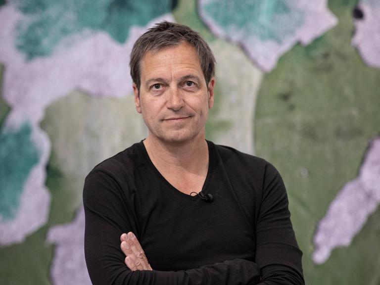 Porträt von Dieter Nuhr mit verschränkten Arnmen vor einem Kunstwerk