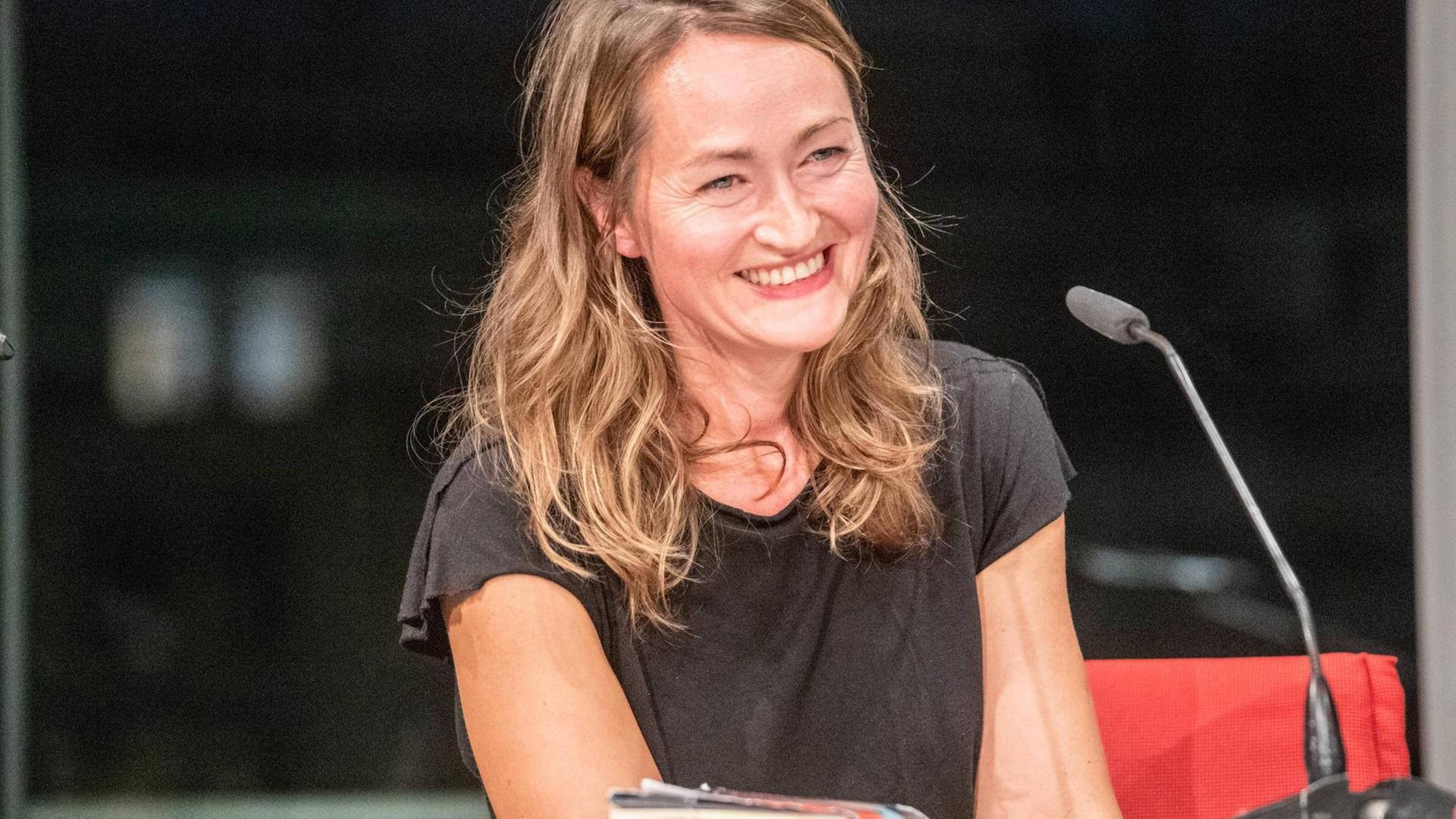 Katerina Poladjan lächelt bei einer Lesung von "Hier sind Löwen" in der Berliner Akademie der Künste ins Publikum. Sie trägt ein dunkles T-Shirt, vor ihr steht ein Mikrofon. Katerina Poladjan lächelt bei einer Lesung von "Hier sind Löwen" in der Berliner Akademie der Künste ins Publikum. Sie trägt ein dunkles T-Shirt, vor ihr steht ein Mikrofon.