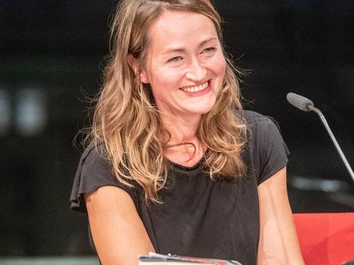 Katerina Poladjan lächelt bei einer Lesung von "Hier sind Löwen" in der Berliner Akademie der Künste ins Publikum. Sie trägt ein dunkles T-Shirt, vor ihr steht ein Mikrofon.