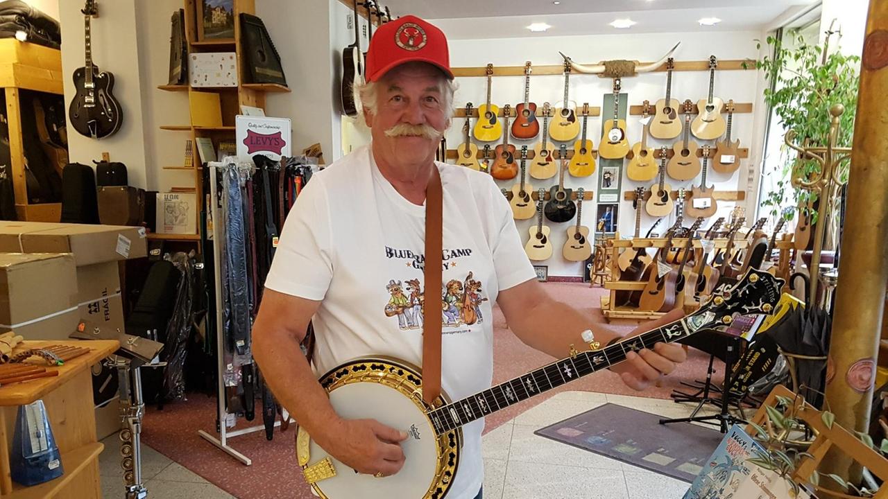 Ein Mann im weißen T-Shirt und roter Kappe halt ein Banjo in der Hand und lächelt in die Kamera. Links neben ihm Gitarren-Gurte, an der Wand hinter ihm hängen Akustik-Gitarren. Ein Mann im weißen T-Shirt und roter Kappe halt ein Banjo in der Hand und lächelt in die Kamera. Links neben ihm Gitarren-Gurte, an der Wand hinter ihm hängen Akustik-Gitarren.