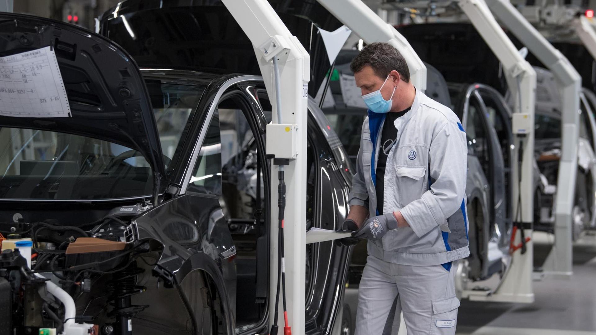 Das Bild zeigt einen Arbeiter an einem Montageband in einer VW-Autofabrik. Der Arbeiter trägt eine Mundnasenmaske. Das Bild zeigt einen Arbeiter an einem Montageband in einer VW-Autofabrik. Der Arbeiter trägt eine Mundnasenmaske.