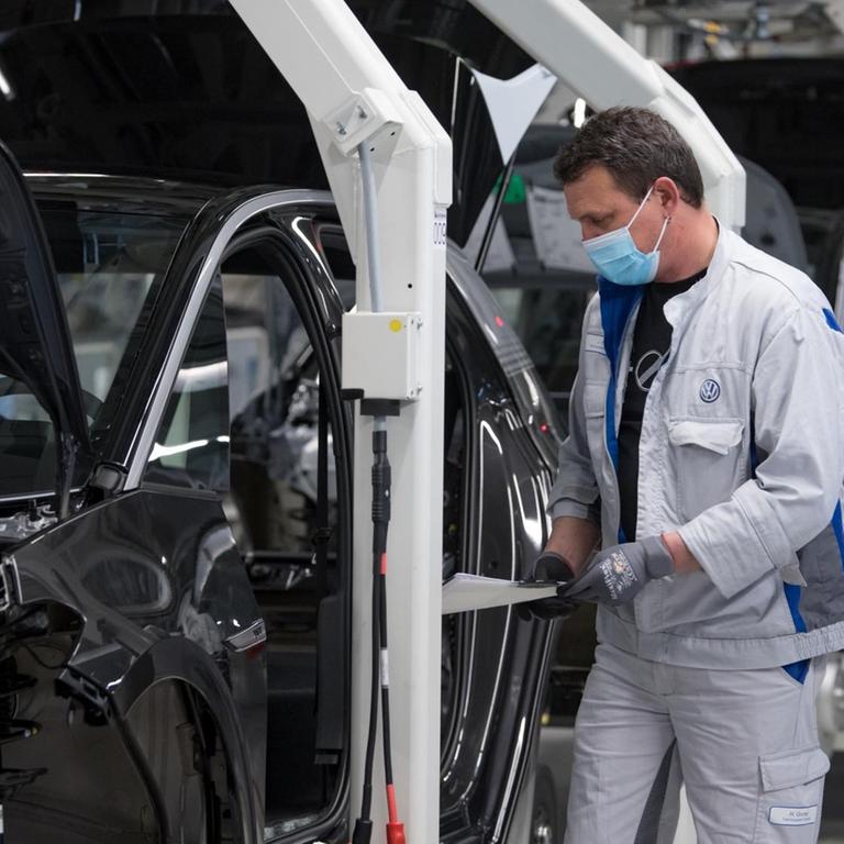 Das Bild zeigt einen Arbeiter an einem Montageband in einer VW-Autofabrik. Der Arbeiter trägt eine Mundnasenmaske.