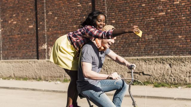 Ein junges Paar auf einem Fahrrad, die Frau steht hinten auf dem Gepäckträger und mach Selfies