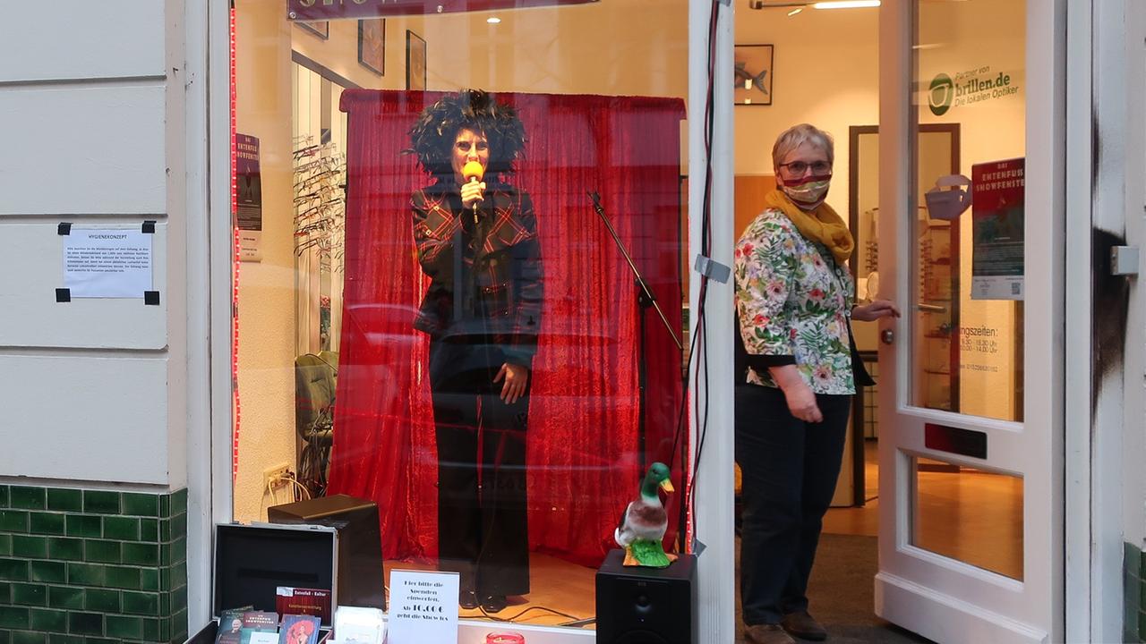 Hinter dem Schaufenster eines Optikers steht eine Schauspielerin vor einem roten Vorhang. Sie trägt eine schwarze Schlaghose und eine auffällige schwarze Perücke. Sie spricht in ein gelbes Mikrofon. Daneben öffnet eine Frau mit Maske die Tür. Hinter dem Schaufenster eines Optikers steht eine Schauspielerin vor einem roten Vorhang. Sie trägt eine schwarze Schlaghose und eine auffällige schwarze Perücke. Sie spricht in ein gelbes Mikrofon. Daneben öffnet eine Frau mit Maske die Tür.