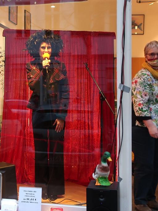 Hinter dem Schaufenster eines Optikers steht eine Schauspielerin vor einem roten Vorhang. Sie trägt eine schwarze Schlaghose und eine auffällige schwarze Perücke. Sie spricht in ein gelbes Mikrofon. Daneben öffnet eine Frau mit Maske die Tür.