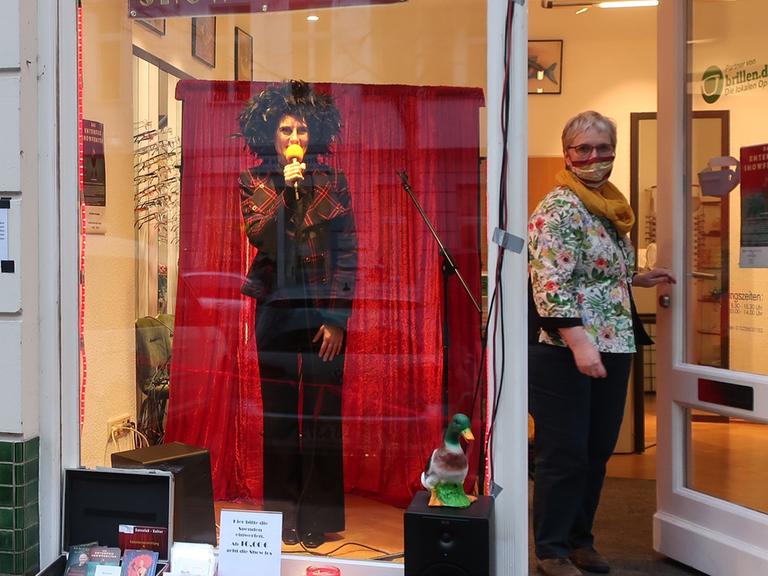 Hinter dem Schaufenster eines Optikers steht eine Schauspielerin vor einem roten Vorhang. Sie trägt eine schwarze Schlaghose und eine auffällige schwarze Perücke. Sie spricht in ein gelbes Mikrofon. Daneben öffnet eine Frau mit Maske die Tür.