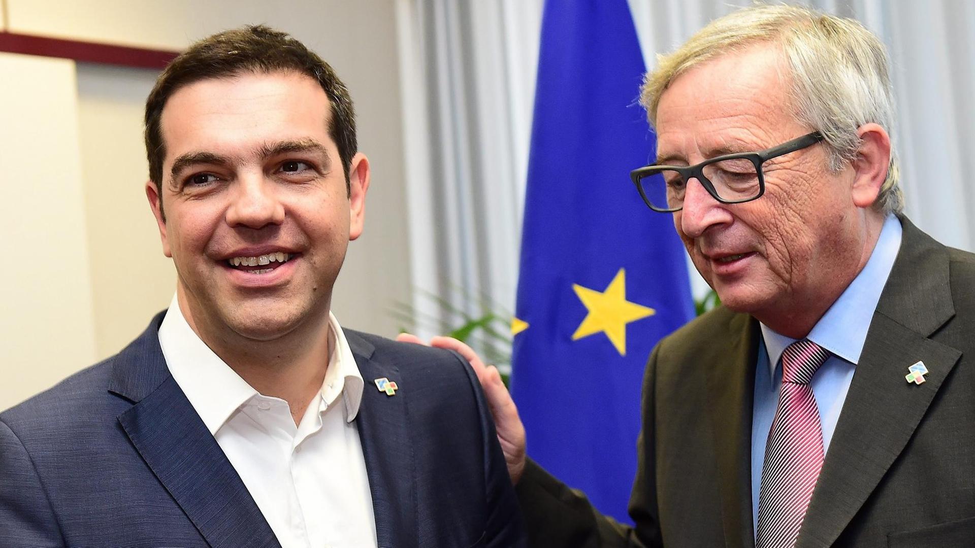 Tsipras und Juncker stehen in einem Raum vor einer EU-Flagge. Tsipras lacht. Juncker hat seine Hand auf Tsipras' Schulter und redet auf ihn ein. Tsipras und Juncker stehen in einem Raum vor einer EU-Flagge. Tsipras lacht. Juncker hat seine Hand auf Tsipras' Schulter und redet auf ihn ein.