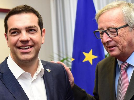 Tsipras und Juncker stehen in einem Raum vor einer EU-Flagge. Tsipras lacht. Juncker hat seine Hand auf Tsipras' Schulter und redet auf ihn ein.