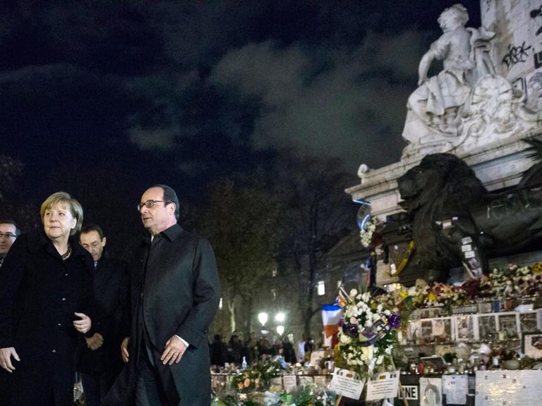Bundeskanzlerin Merkel und Frankreichs Präsident Hollande auf der Place de la République in Paris, es ist dunkel, beide tragen Mäntel.