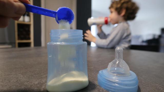 In eine Plastikflasche wird Milchpulver eingefüllt. Im Hintergrund trinkt ein Kleinkind seine Milch.