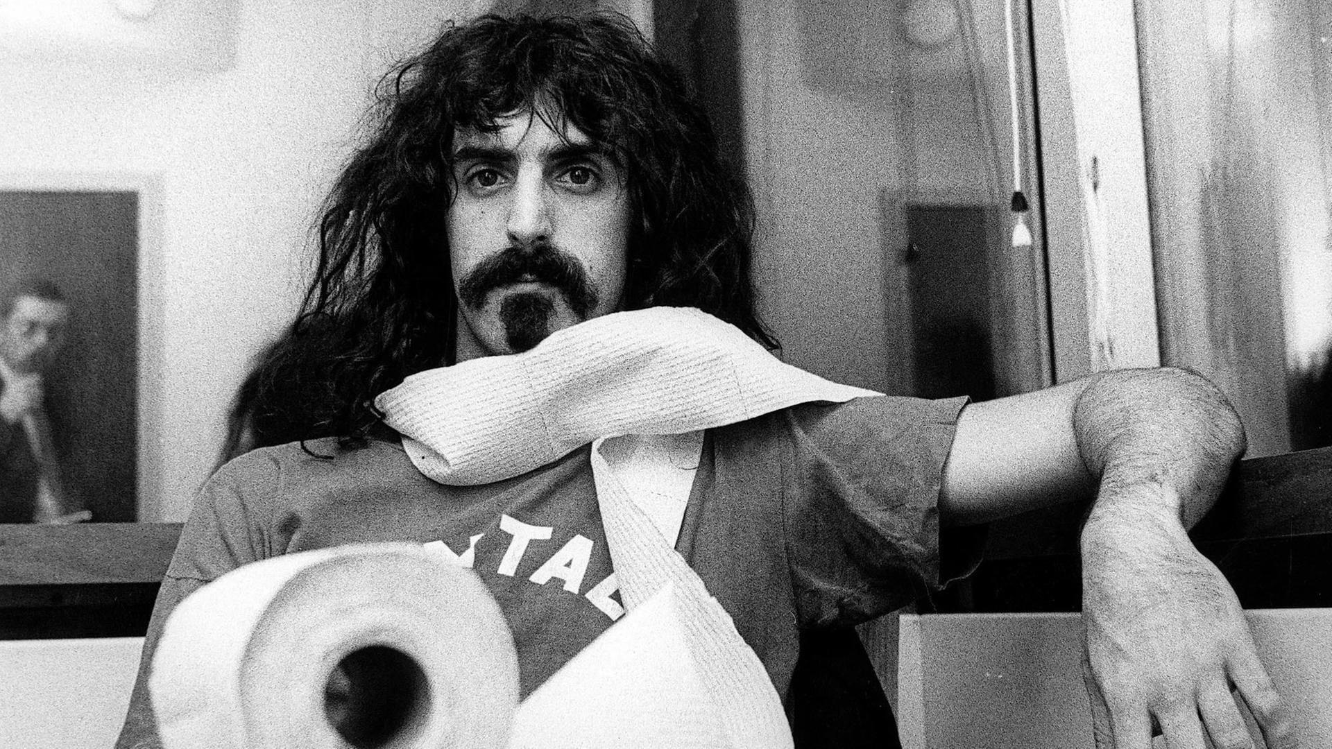 Porträt von Frank Zappa an einem Tisch mit einer Klorolle, die er sich um den Hals gewickelt hat. Porträt von Frank Zappa an einem Tisch mit einer Klorolle, die er sich um den Hals gewickelt hat.