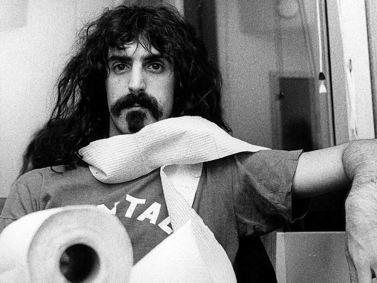 Porträt von Frank Zappa an einem Tisch mit einer Klorolle, die er sich um den Hals gewickelt hat.