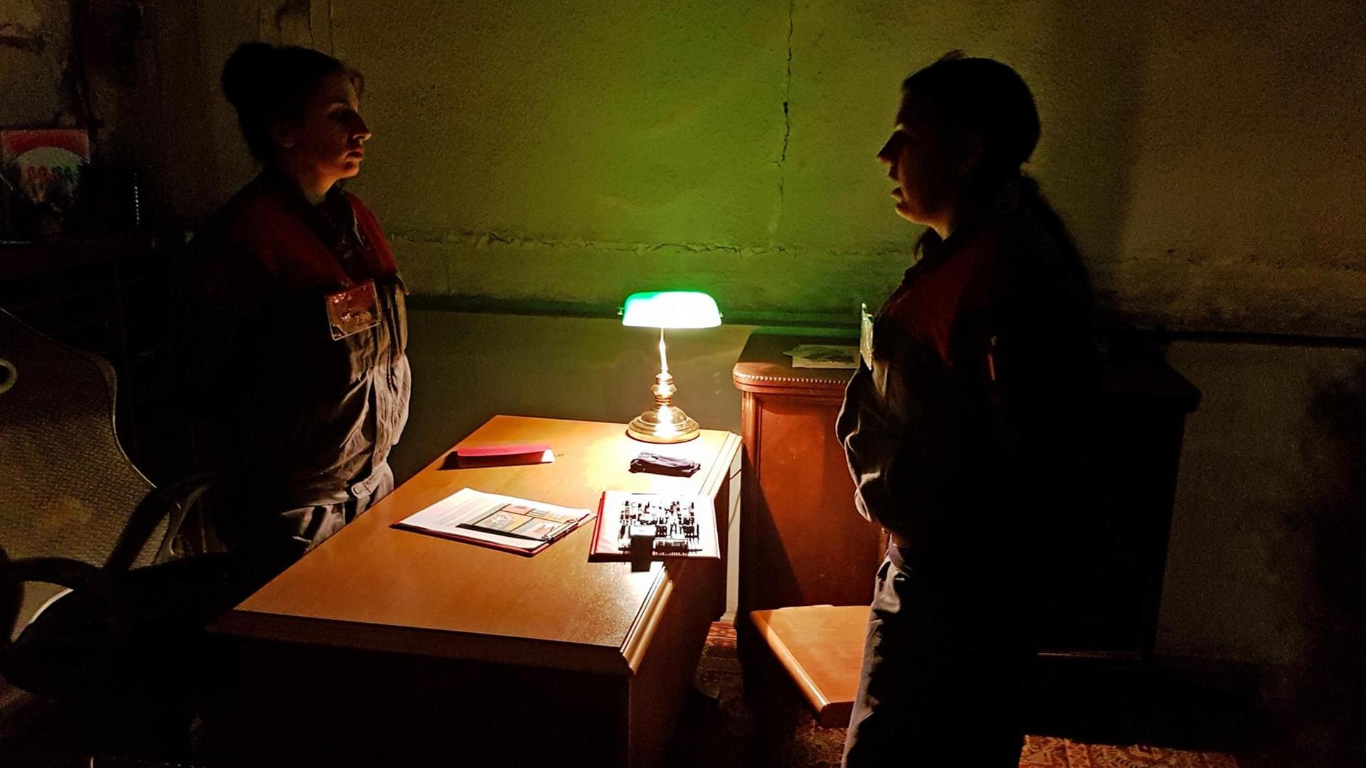 Szene aus dem Rollenspiel "Inside": In einem dunklem Büro stehen sich zwei Frauen in Uniformen gegenüber, zwischen ihnen ein Schreibtisch. Szene aus dem Rollenspiel "Inside": In einem dunklem Büro stehen sich zwei Frauen in Uniformen gegenüber, zwischen ihnen ein Schreibtisch.