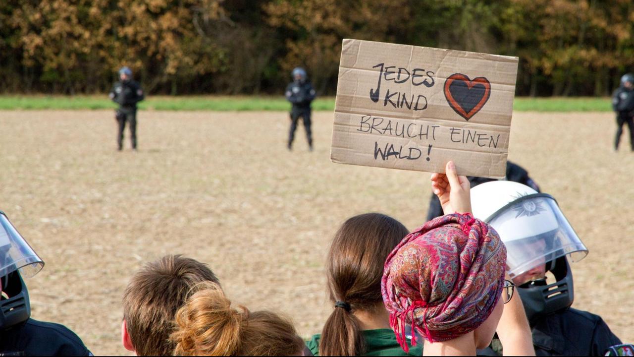 Demonstranten stehen vor Polizisten. Im Hintergrund ist ein Wald zu sehen. Eine Demonstrantin hält ein Schild mit der Aufschirft "Jedes Kind braucht einen Wald" hoch. Demonstranten stehen vor Polizisten. Im Hintergrund ist ein Wald zu sehen. Eine Demonstrantin hält ein Schild mit der Aufschirft "Jedes Kind braucht einen Wald" hoch.