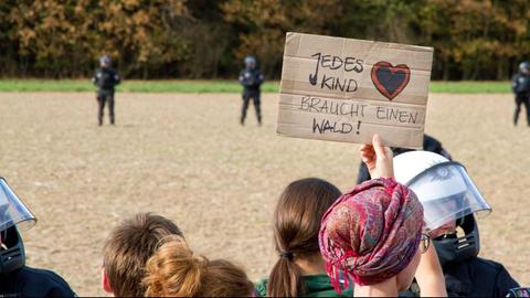 Demonstranten stehen vor Polizisten. Im Hintergrund ist ein Wald zu sehen. Eine Demonstrantin hält ein Schild mit der Aufschirft "Jedes Kind braucht einen Wald" hoch.