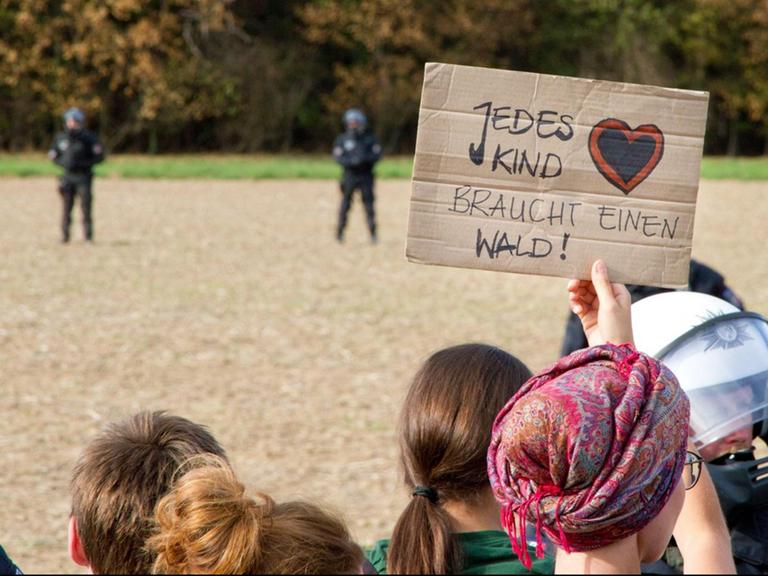 Demonstranten stehen vor Polizisten. Im Hintergrund ist ein Wald zu sehen. Eine Demonstrantin hält ein Schild mit der Aufschirft "Jedes Kind braucht einen Wald" hoch.