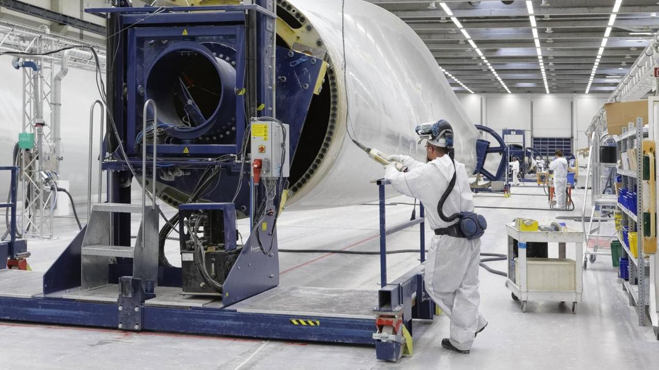 Blick in die Werkhalle. Arbeiter in Schutzanzügen stehen an einem großen Flügel einer Windkraftanlage. Blick in die Werkhalle. Arbeiter in Schutzanzügen stehen an einem großen Flügel einer Windkraftanlage.