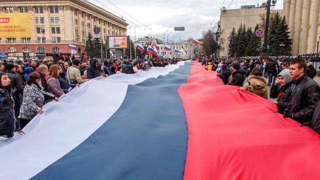 Eine Russlandflagge erstreckt sich hunderte Meter weit. An der Seite stehen Demonstranten und halten sie hoch.