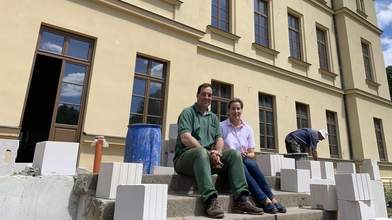Verena und Till-Alexander Herzer sitzen auf der Treppe vor ihrem Haus. Er trägt grüne, dreckige Arbeitskleidung. Verena und Till-Alexander Herzer sitzen auf der Treppe vor ihrem Haus. Er trägt grüne, dreckige Arbeitskleidung.