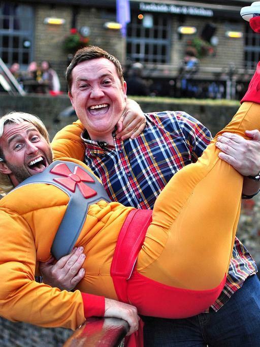 Zwei Männer lachen in die Kamera. Einer trägt ein Superhelden-Kostüm und eine blonde Perücke, der andere trägt ihn auf dem Arm.