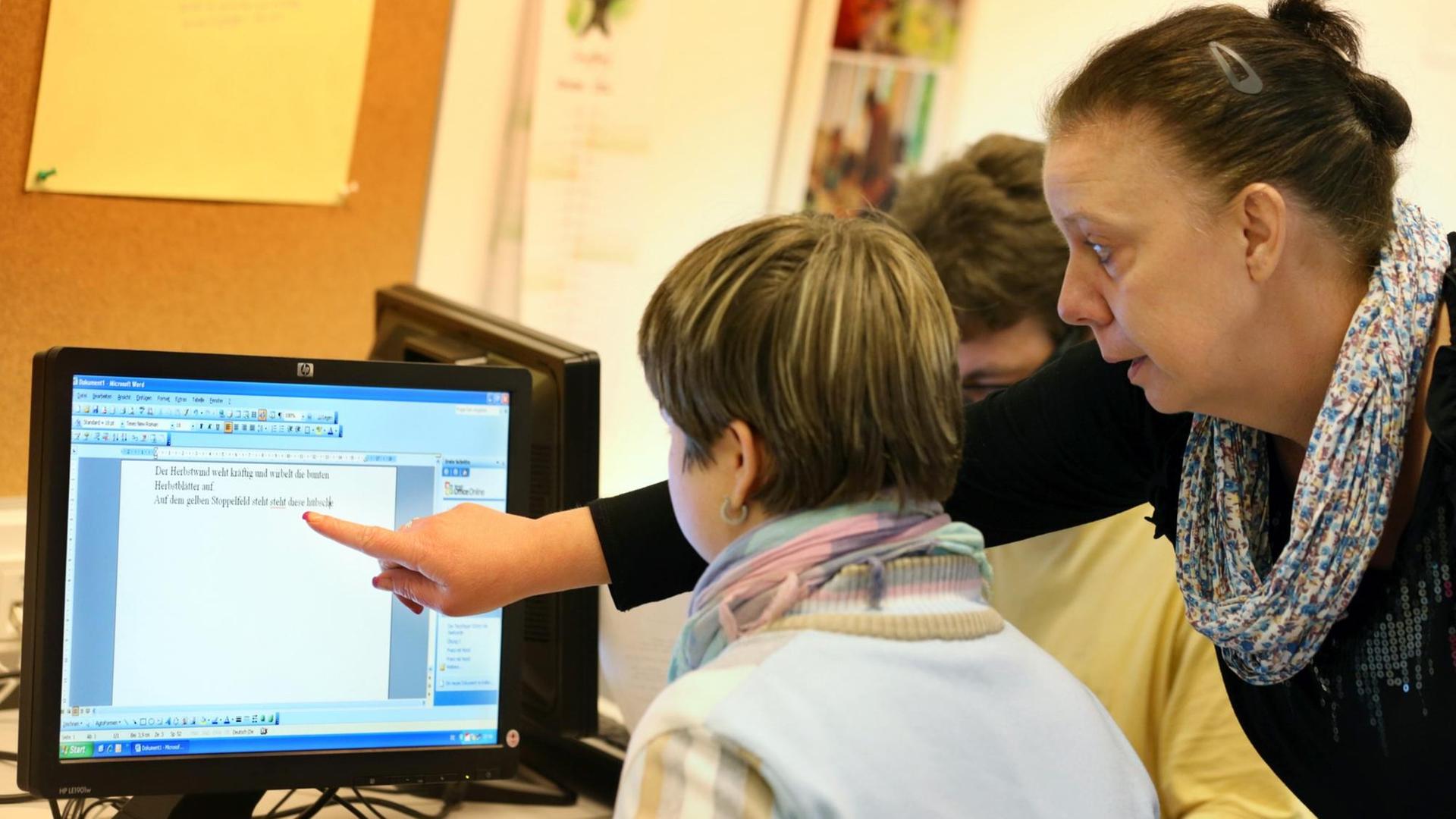 Eine Frau sitzt an einem Computer und schreibt etwas, eine andere Frau steht hinter ihr und zeigt auf etwas auf dem Bildschirm. Eine Frau sitzt an einem Computer und schreibt etwas, eine andere Frau steht hinter ihr und zeigt auf etwas auf dem Bildschirm.