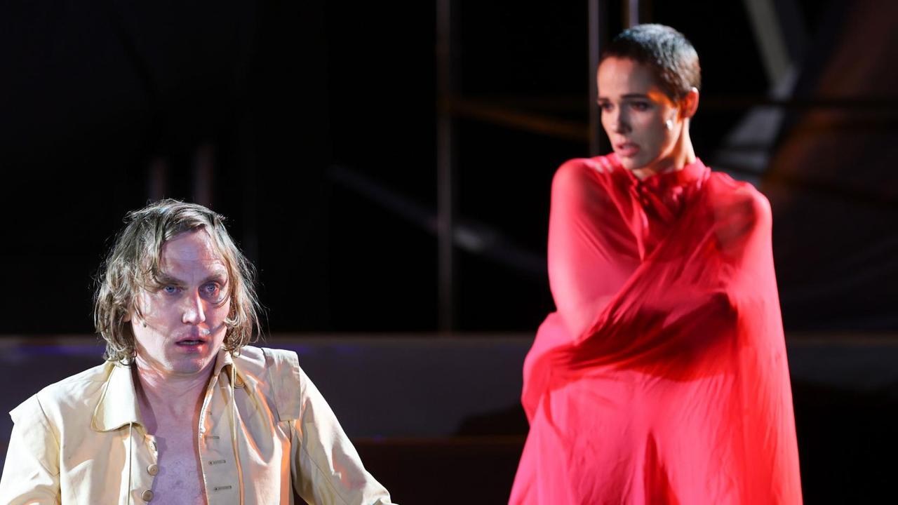 Zwei Schauspieler sind auf einer Bühne. Im Vordergrund ein Mann mit gelber Jacke, im Hintergrund eine Frau in rotem Gewandt.