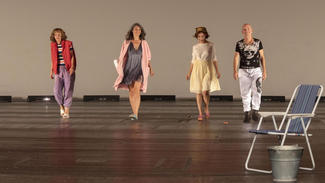 Drei Frauen und ein Mann gehen nebeneinander auf der Bühne. Die Frauen tragen pastellfarbene, bunte Kleidung und Badelatschen, der Mann eine weiße Hose und ein dunkles T-Shirt, auf dem in weiß ein Totenkopf zu sehen ist. Am rechten Rand steht vor den vier Personen ein aufgeklappter Camping-Stuhl. Drei Frauen und ein Mann gehen nebeneinander auf der Bühne. Die Frauen tragen pastellfarbene, bunte Kleidung und Badelatschen, der Mann eine weiße Hose und ein dunkles T-Shirt, auf dem in weiß ein Totenkopf zu sehen ist. Am rechten Rand steht vor den vier Personen ein aufgeklappter Camping-Stuhl.