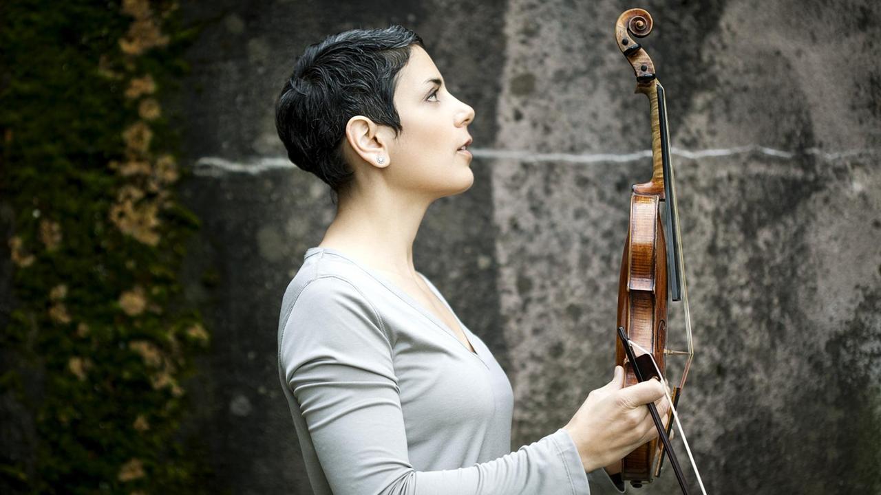 Die Geigerin Leila Schayegh im Profil steht vor einer Steinwand und hält ihren Bogen und ihre Violine aufrecht vor sich Die Geigerin Leila Schayegh im Profil steht vor einer Steinwand und hält ihren Bogen und ihre Violine aufrecht vor sich