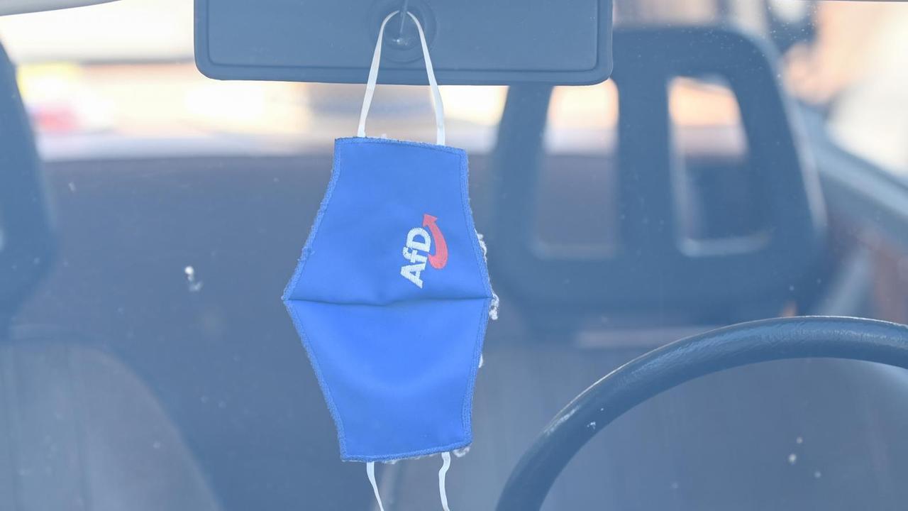 Eine Mund-Nasen-Bedeckung mit dem Logo der AfD hängt am Rande einer Wahlkampfveranstaltung am Rückspiegel eines Trabants.