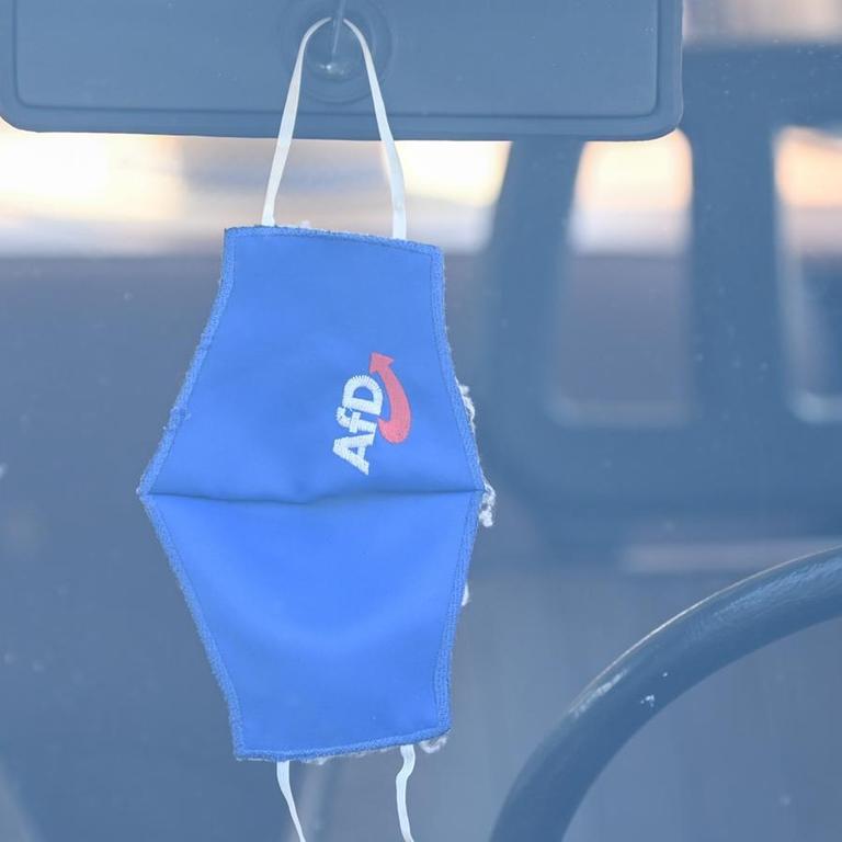 Eine Mund-Nasen-Bedeckung mit dem Logo der AfD hängt am Rande einer Wahlkampfveranstaltung am Rückspiegel eines Trabants.