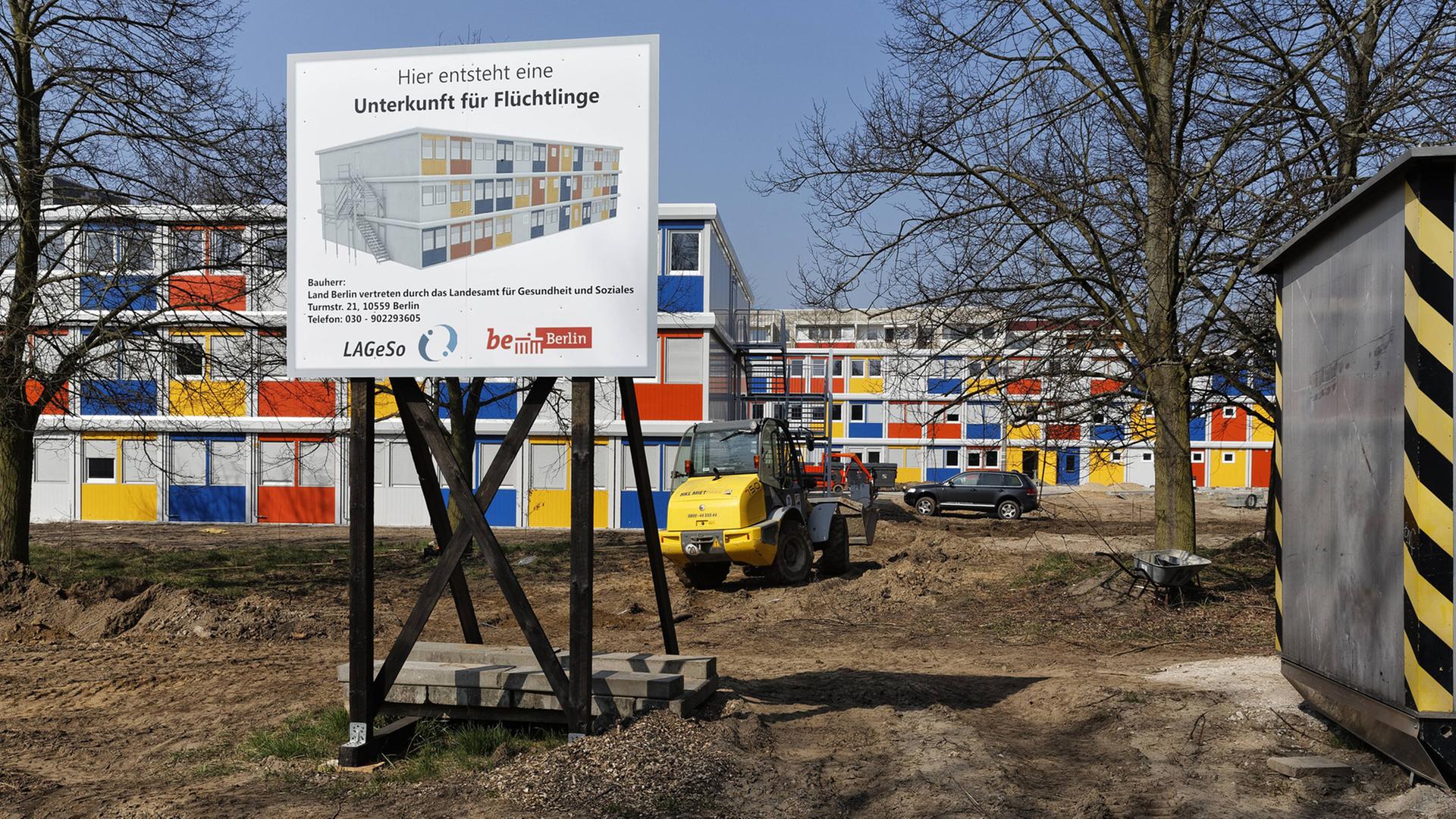 Neben- und übereinandergestapelte Container in blau, rot, gelb und weiß auf einer Baustelle, davor ein Schild mit dem Text "Hier entsteht eine Unterkunft für Flüchtlinge". Neben- und übereinandergestapelte Container in blau, rot, gelb und weiß auf einer Baustelle, davor ein Schild mit dem Text "Hier entsteht eine Unterkunft für Flüchtlinge".