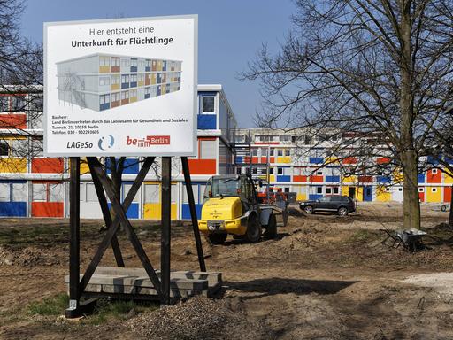 Neben- und übereinandergestapelte Container in blau, rot, gelb und weiß auf einer Baustelle, davor ein Schild mit dem Text "Hier entsteht eine Unterkunft für Flüchtlinge".