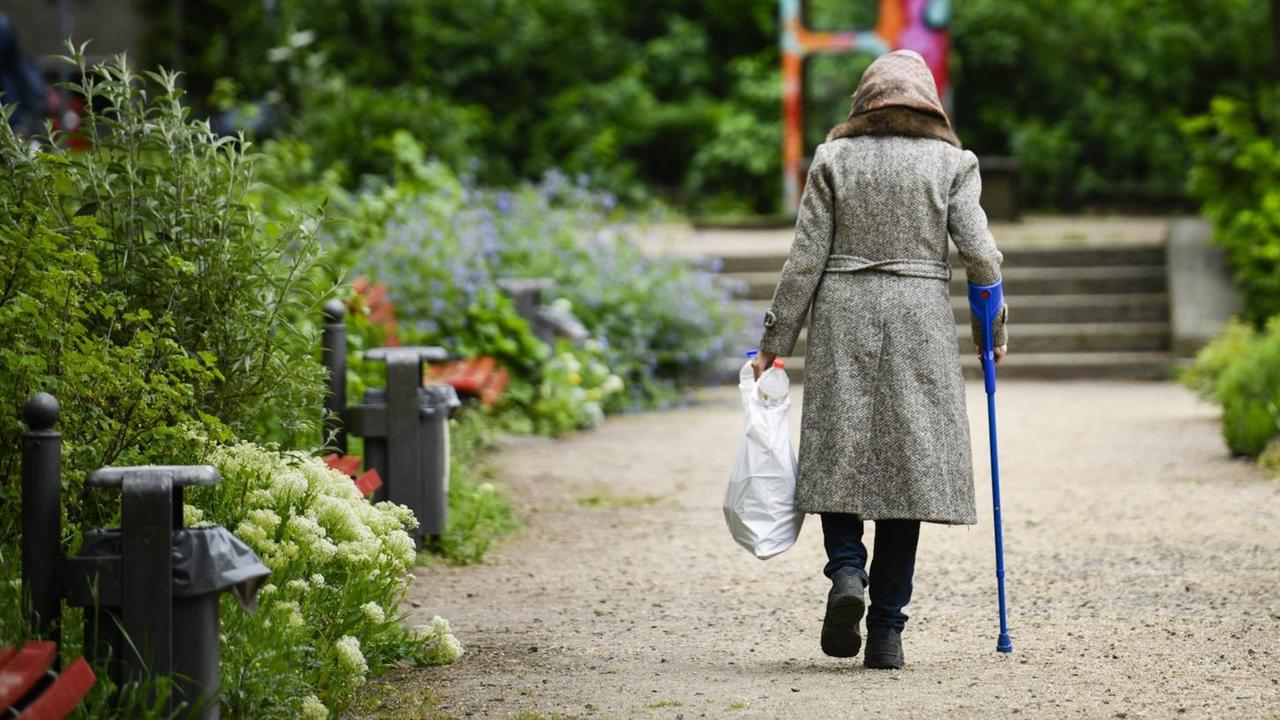 Rückansicht einer Rentnerin mit Krücke, die durch einen Park geht und dabei eine Plastiktüte mit Pfandflaschen trägt. Rückansicht einer Rentnerin mit Krücke, die durch einen Park geht und dabei eine Plastiktüte mit Pfandflaschen trägt.