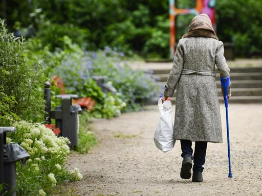 Rückansicht einer Rentnerin mit Krücke, die durch einen Park geht und dabei eine Plastiktüte mit Pfandflaschen trägt.