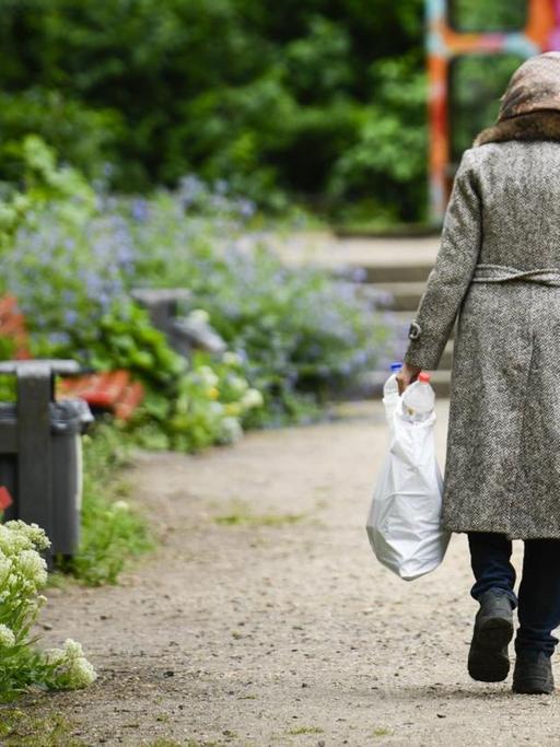 Rückansicht einer Rentnerin mit Krücke, die durch einen Park geht und dabei eine Plastiktüte mit Pfandflaschen trägt.