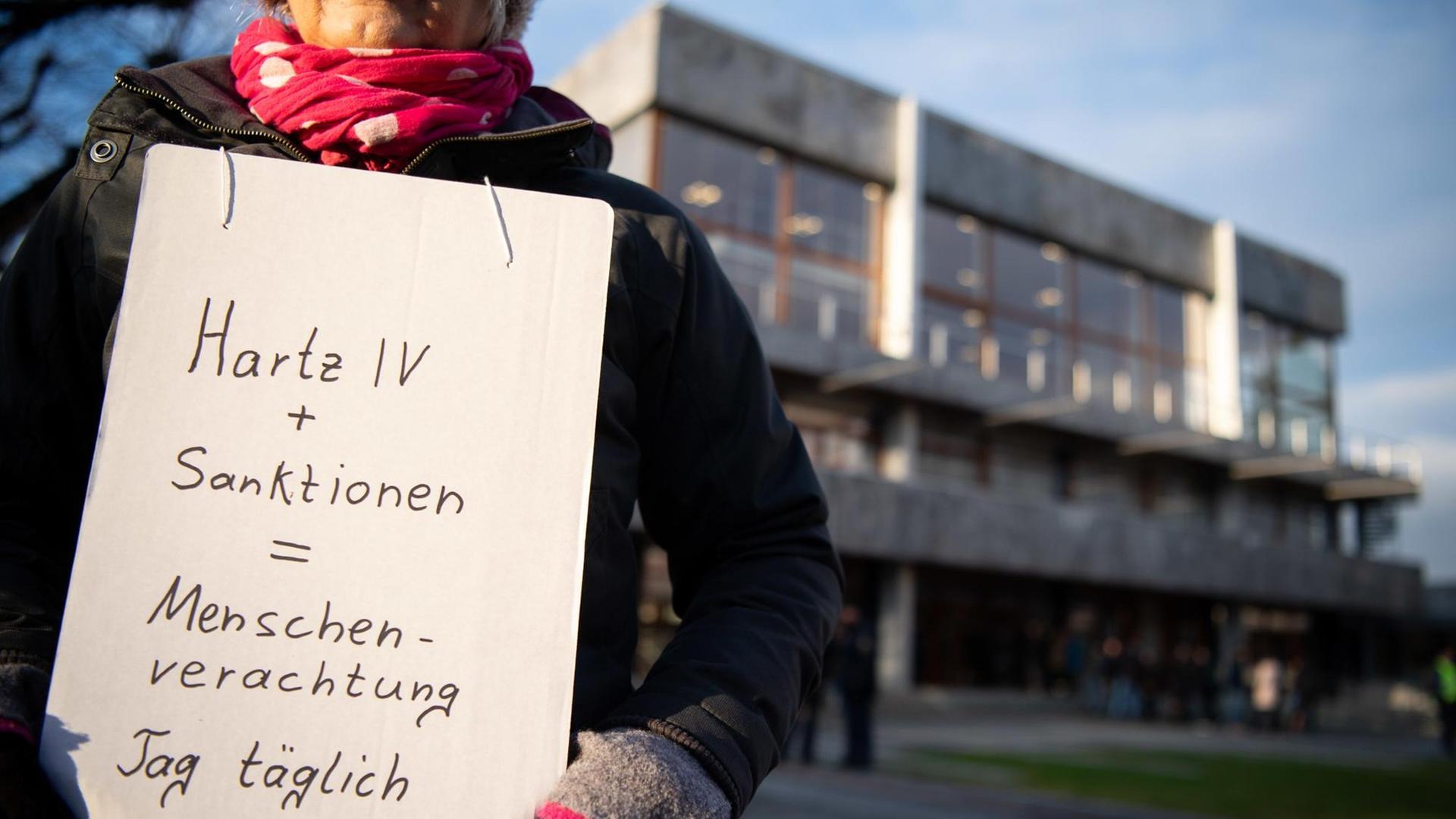 Eine Frau steht mit einem Schild während der mündlichen Verhandlung über die Rechtmäßigkeit von Hartz-IV-Sanktionen vor dem Bundesverfassungsgericht. Das Sozialgericht Gotha hält die Praxis, nach der Hartz-IV-Empfängern Leistungskürzungen drohen, wenn sie ihren Pflichten nicht nachkommen, für verfassungswidrig. Daher hat es einen Fall in Karlsruhe vorgelegt. Das Urteil wird in der Regel einige Monate nach der Verhandlung verkündet. Foto: Sebastian Gollnow/dpa | Verwendung weltweit Eine Frau steht mit einem Schild während der mündlichen Verhandlung über die Rechtmäßigkeit von Hartz-IV-Sanktionen vor dem Bundesverfassungsgericht. Das Sozialgericht Gotha hält die Praxis, nach der Hartz-IV-Empfängern Leistungskürzungen drohen, wenn sie ihren Pflichten nicht nachkommen, für verfassungswidrig. Daher hat es einen Fall in Karlsruhe vorgelegt. Das Urteil wird in der Regel einige Monate nach der Verhandlung verkündet. Foto: Sebastian Gollnow/dpa | Verwendung weltweit