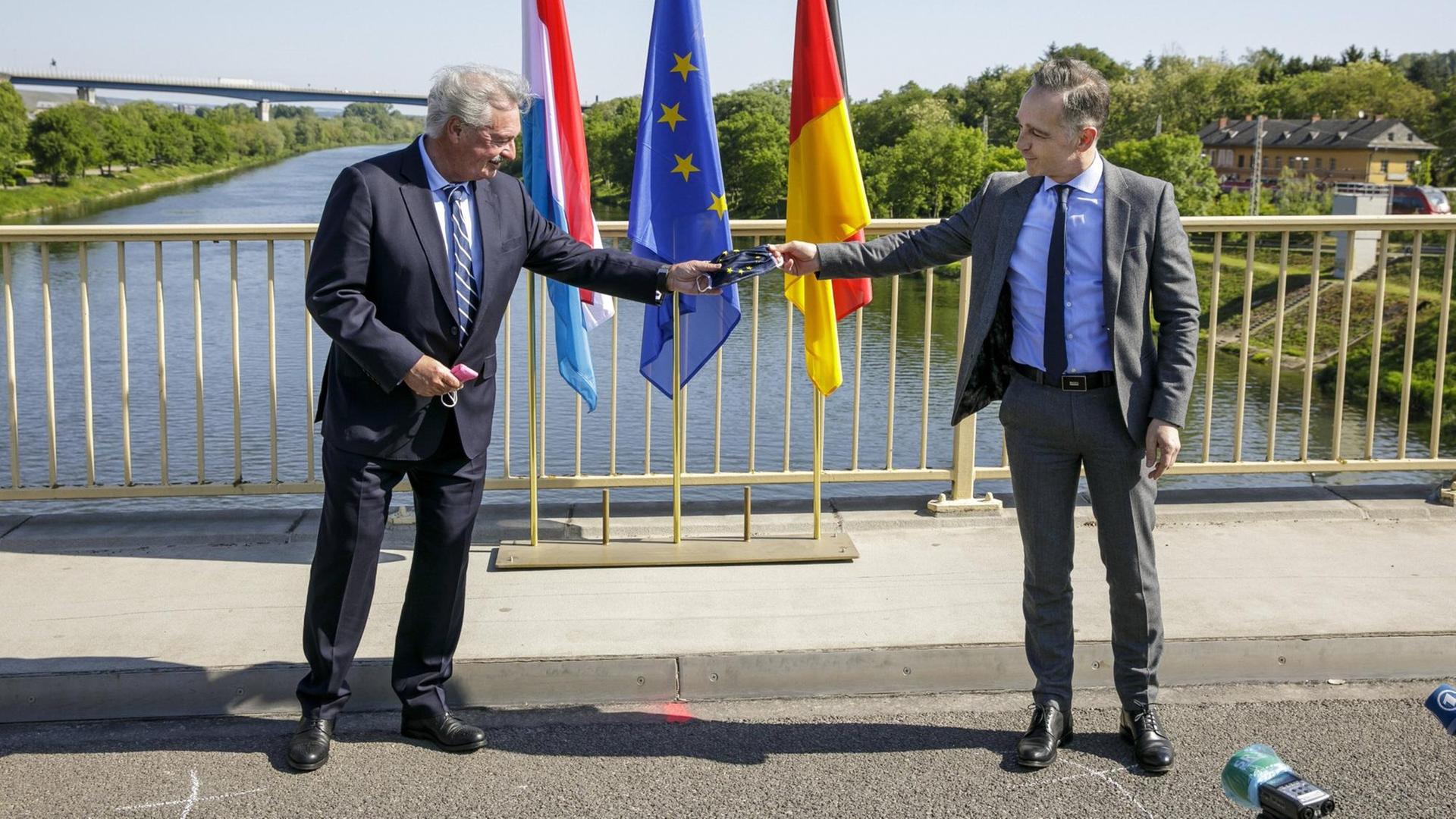 Heiko Maas und Jean Asselborn stehen auf einer Brücke der deutsch-luxemburgischen Grenze und halten als symbolische Geste eine Corona-Gesichtsmaske mit dem Wappen der Europäischen Union.
