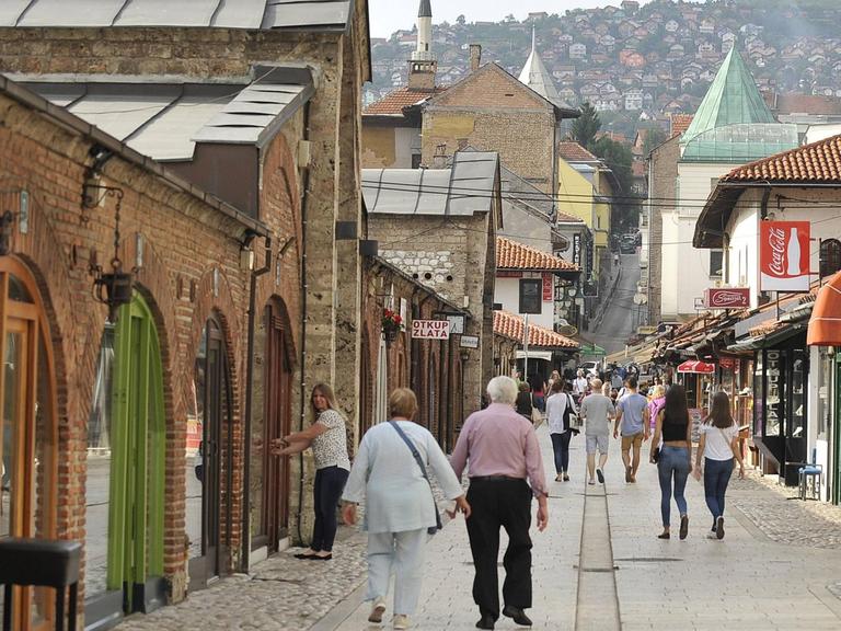 Eine Fußgänger-Straße in der Altstadt Sarajevos mit Blick auf die bewohnten Hügel im Hintergrund. Links ein altes Backsteingebäude mit Torbögen. Aufgenommen am 22.06.2016