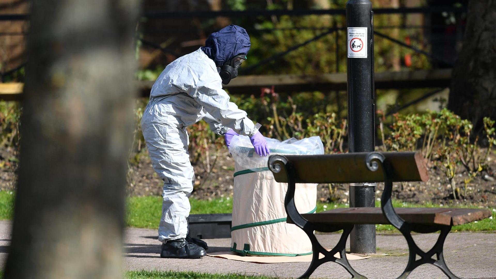 Ein Mann in weißer Schutzkleidung und Atemmaske greift einen Mülleimer in einer Grünanlage. Ein Mann in weißer Schutzkleidung und Atemmaske greift einen Mülleimer in einer Grünanlage.