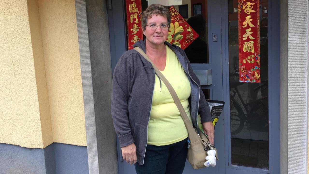 Elke Raue steht am Eingang eines Hauses in Oak Garden. Tücher mit chinesischen Schriftzeichen zieren die Tür. Raue trägt ein gelbes Shirt, graue Jacke und eine Umhängetasche.  