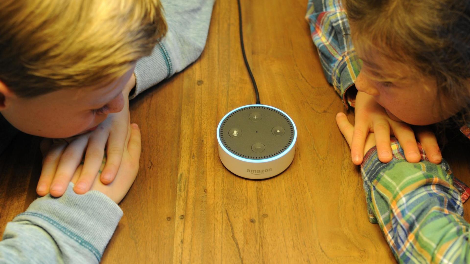 Der Amazon Echo Dot ist ein Lautsprecher, der auf den Namen "Alexa" hört und als Sprachschnittstelle zu Amazon-Produkten fungiert. Er steht auf einem Tisch. Zwei Kinder lehnen sich mit Händen und Köpfen auf den Tisch und schauen den Lautsprecher an.