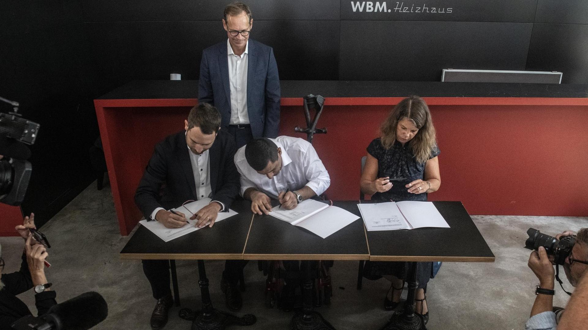 02.08.2019, Berlin: Rahim Nagibulla unterzeichnet den ersten Arbeitsvertrag im Rahmen des bundesweit einmaligen Projekts "Solidarisches Grundeinkommen". Gegengezeichnet wird sein Arbeitsvertrag von Christina Geib (r) und Jan Robert Kowalewski (l) von der Wohnungsbaugesellschaft Mitte. Der Regierende Bürgermeister Michael Müller (SPD) verfolgt das Geschehen. 02.08.2019, Berlin: Rahim Nagibulla unterzeichnet den ersten Arbeitsvertrag im Rahmen des bundesweit einmaligen Projekts "Solidarisches Grundeinkommen". Gegengezeichnet wird sein Arbeitsvertrag von Christina Geib (r) und Jan Robert Kowalewski (l) von der Wohnungsbaugesellschaft Mitte. Der Regierende Bürgermeister Michael Müller (SPD) verfolgt das Geschehen.